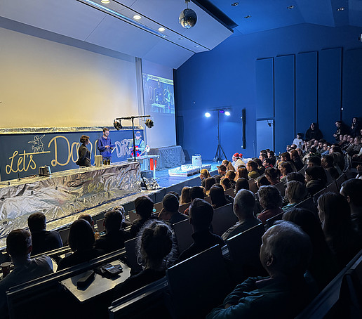 Full rows in the Audimax. Elaborate experimental equipment is set up on the podium.
