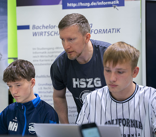 HSZG-Mitarbeiter informiert zwei sitzende Schüler am Laptop.
