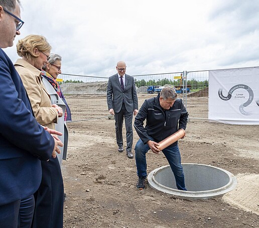 A man stands with one leg in a hole dug out of the ground and holds the foundation stone in his hands. The participants stand around him.