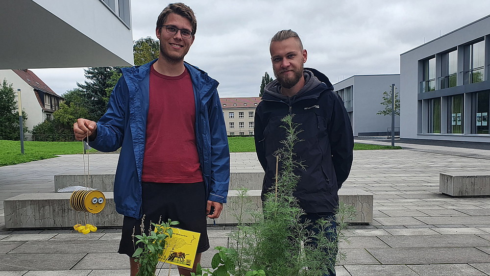 Zwei junge Männer stehen auf dem Zittauer Campus. Einer hält ein Bienenhotel in der Hand.
