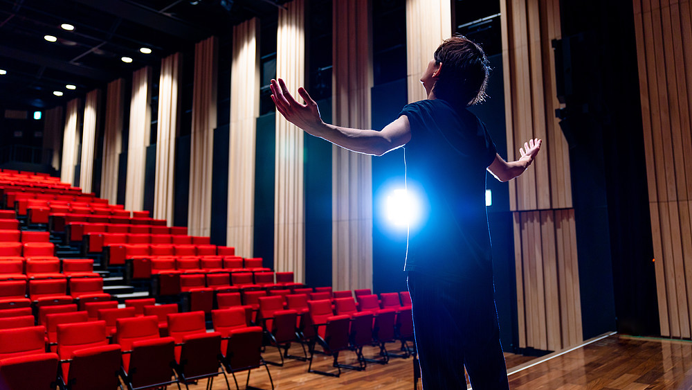 Ein junger Mann steht auf einer Theaterbühne, leeren rotgepolsterten Sitzreihen zugewandt.