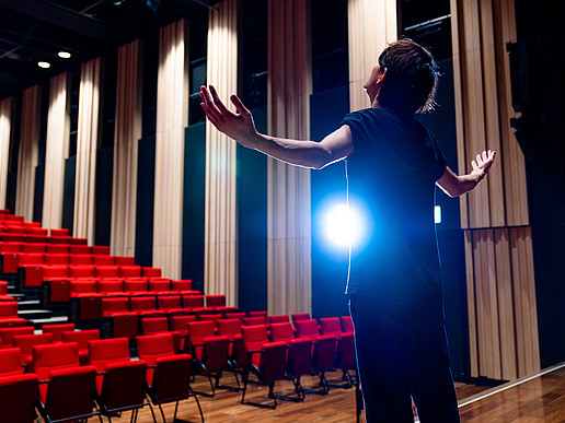 Ein junger Mann steht auf einer Theaterbühne, leeren rotgepolsterten Sitzreihen zugewandt.