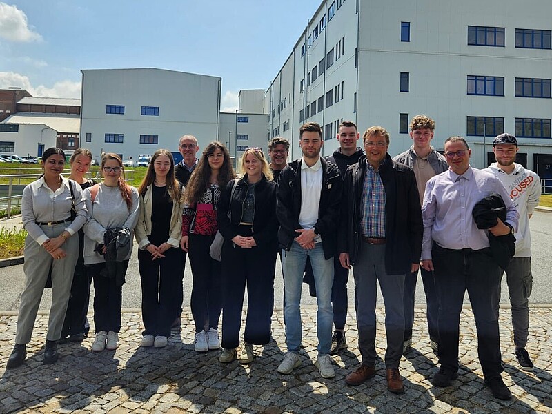 A group of students and lecturers stand on a square in front of the fit GmbH production buildings.