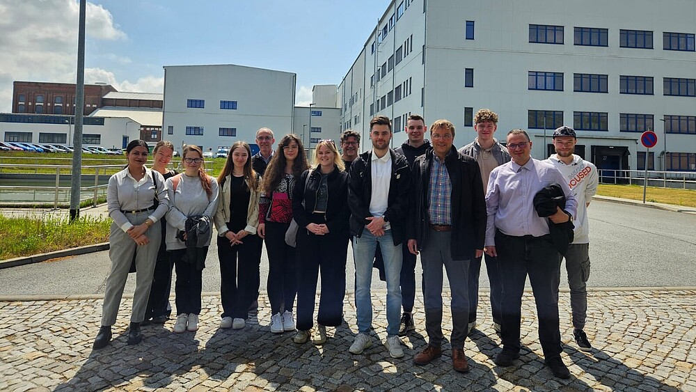 A group of students and lecturers stand on a square in front of the fit GmbH production buildings.