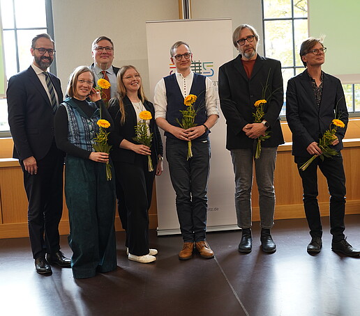 Seven people stand next to each other, holding flowers in their hands and looking into the camera.