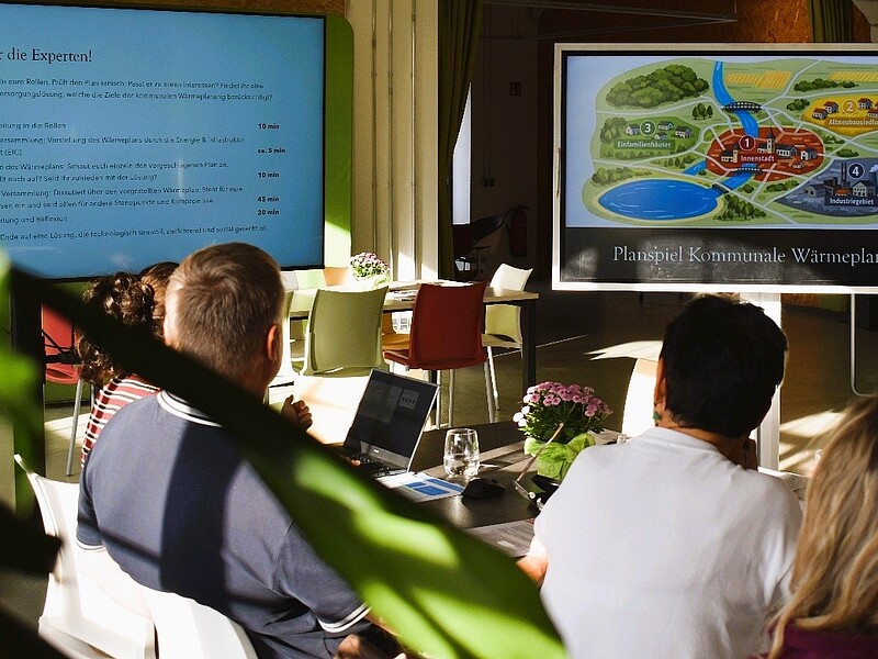 Workshop participants are seated at a long table and looking at a screen.
