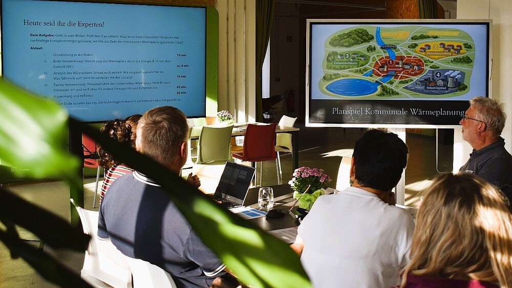 Workshop participants are seated at a long table and looking at a screen.