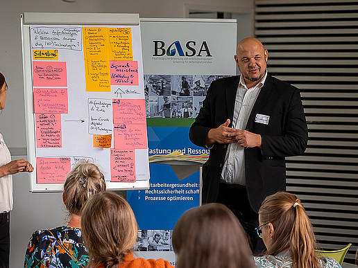 A professor stands in front of full rows of seats and explains something using a board covered with slips of paper.