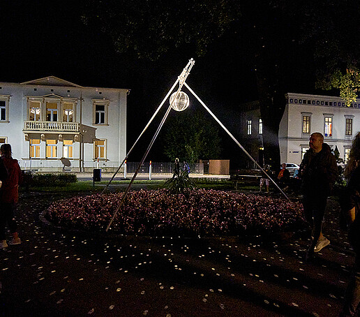 A disco ball hangs from a tube suspension and is illuminated. Visitors walk past it.