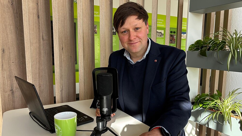 Daniel Winkler sits in front of a microphone and laptop at a high table and smiles into the camera.