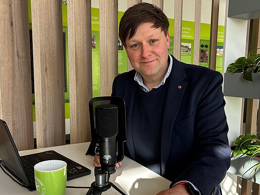 Daniel Winkler sitzt vor einem Mikrofon und Laptop an einem Stehtisch und lächelt in die Kamera.