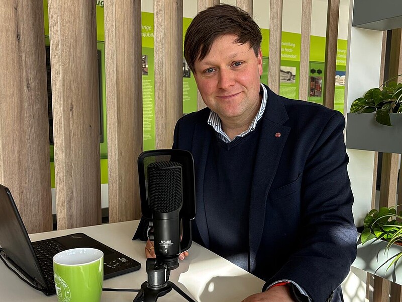 Daniel Winkler sits in front of a microphone and laptop at a high table and smiles into the camera.