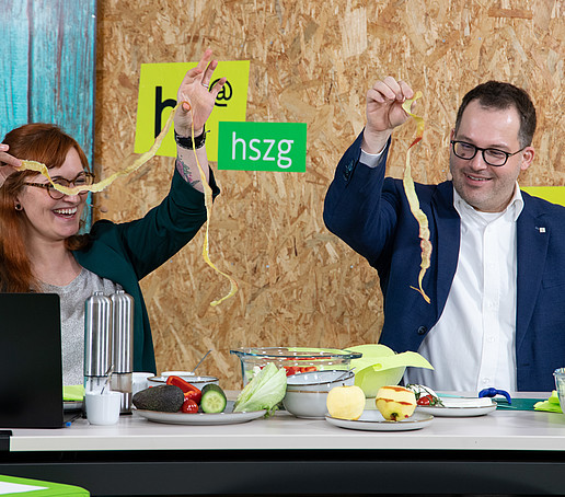 Victoria Hohlfeld und der Rektor stehen am Tisch im Aufnahmestudio und halten jeweils ihre abgeschnittene Apfelschale in die Höhe zum direkten Längenvergleich.