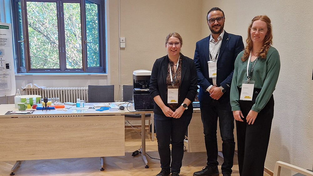 Prof. Dr. Elisa Wirthgen, Ali Al Hamad und Pia Schubert stehen vor dem HSZG-Stand mit Mikroskop und Informationsmaterialien zur biomedizinischen Forschung.