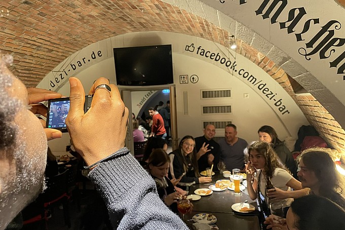 Eine Gruppe von Kollegiaten sitzt an einem Tisch in der Dezi Bar und hat einen fröhlichen Abend. Ein Teilnehmer hält eine Kamera, um die Erinnerungen festzuhalten, während andere Gäste im Hintergrund entspannt zusammensitzen und essen.