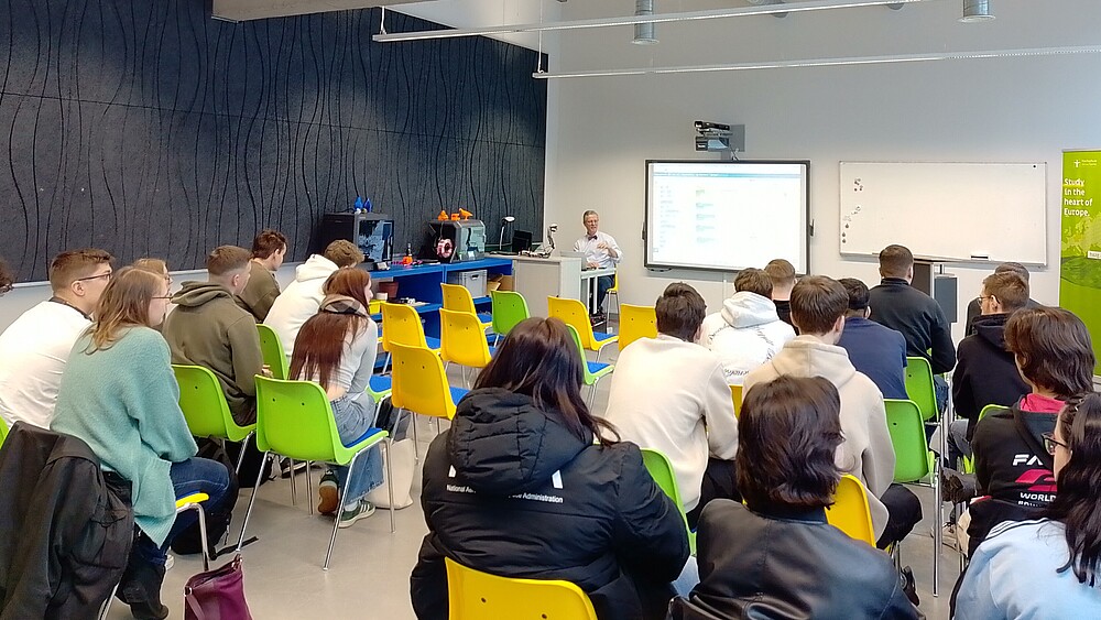 Pupils sit on rows of chairs and listen to the speaker, who stands in front of them next to a screen.