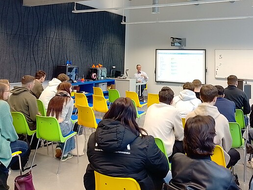 Pupils sit on rows of chairs and listen to the speaker, who stands in front of them next to a screen.