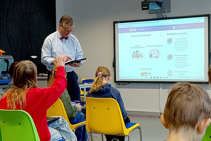 Prof. ten Hagen stands next to a screen and looks at a tablet. Primary school pupils sit in rows in front of him. One pupil raises her arm.
