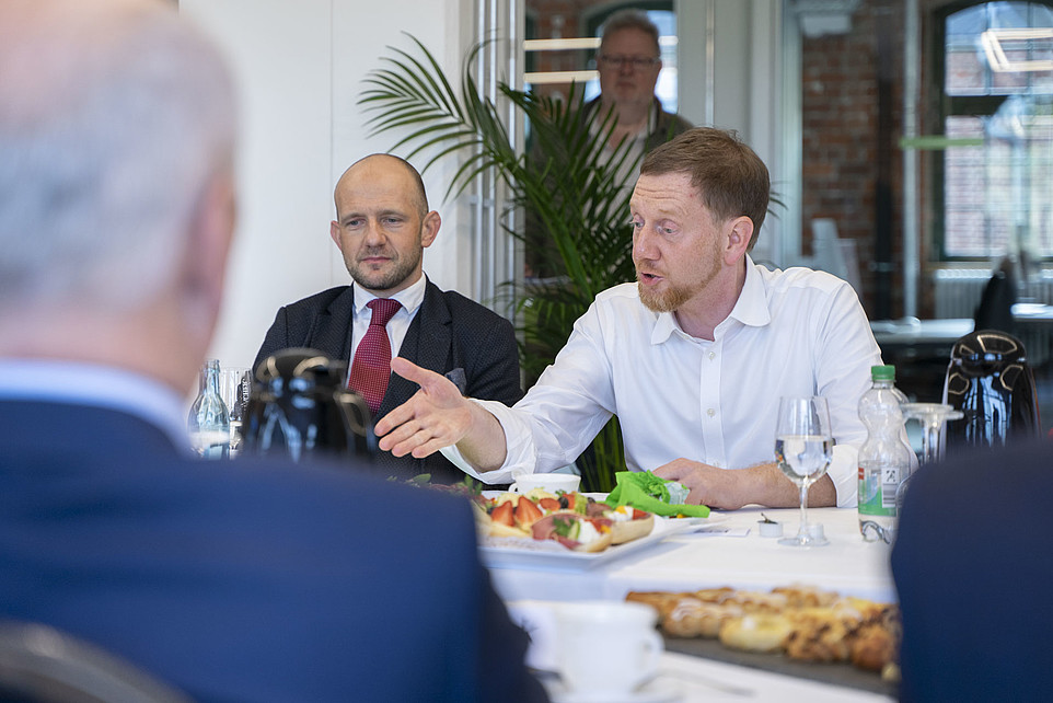 Landrat Meyer und Ministerpräsident Kretschmer im Gespräch sitzen an einem Konferenztisch, Kretschmer gestikuliert erläuternd, im Vordergrund ist der Rücken einer weiteren Person zu sehen.