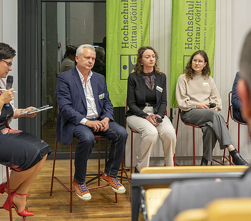 Talk guests sit next to each other on high chairs in the panel, the presenter speaks into a microphone.