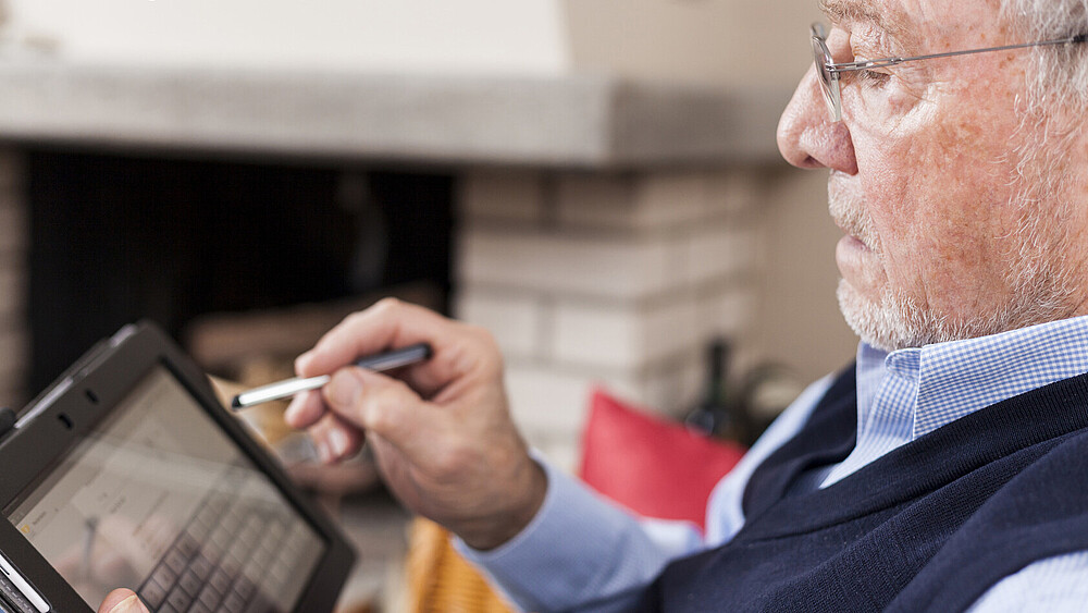 An elderly man wearing glasses is holding a tablet in one hand and a stylus in the other, which he uses to navigate the screen.