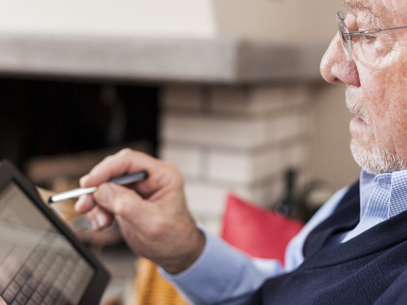 An elderly man wearing glasses is holding a tablet in one hand and a stylus in the other, which he uses to navigate the screen.