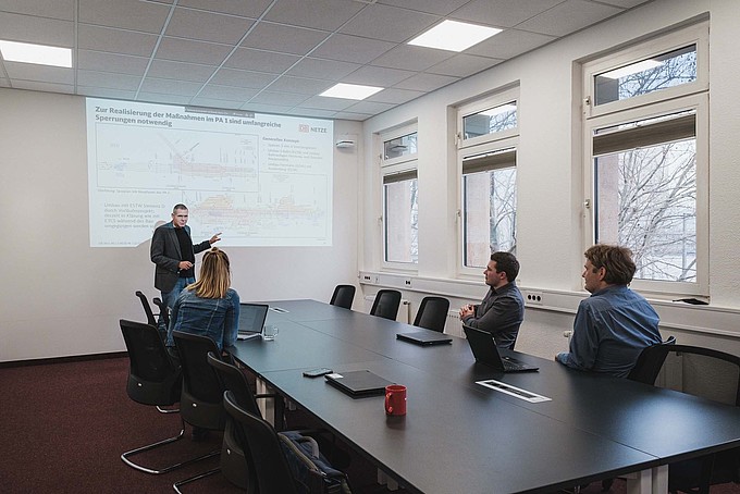 In einem Konferenzraum hält ein Mann eine Präsentation vor drei Zuhörenden.