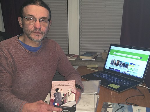 Steffen Seifert holds his book up to the camera and sits at his desk.
