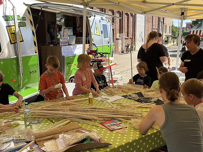Children and a supervisor are sitting at a large table, with natural fibers on the table in front of them that they are using to make things. In the background is the university's mobile learning center trailer.