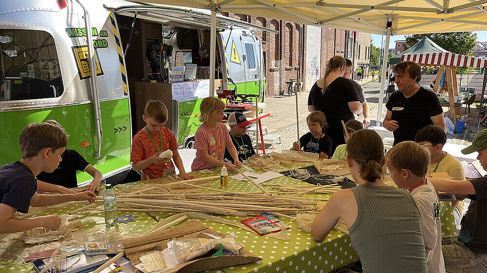Children and a supervisor are sitting at a large table, with natural fibers on the table in front of them that they are using to make things. In the background is the university's mobile learning center trailer.