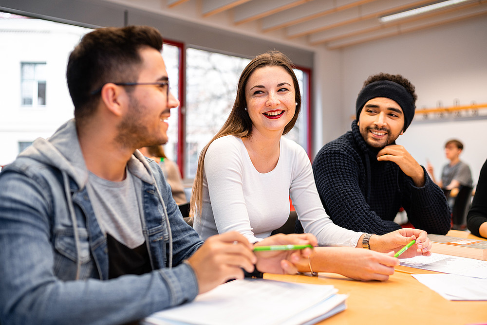 Studierende mit Migrationshintergrund sitzen an einem Schreibtisch lachend und unterhalten sich.