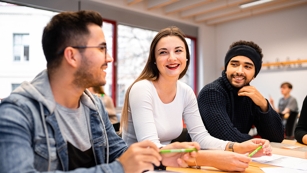 Drei Studierende sitzen am Tisch, unterhalten sich und lachen dabei.