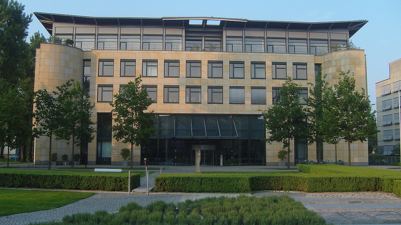 4-storey light brown building with large window front with 6 green deciduous trees and neatly trimmed small hedges framing the sidewalks in front of it