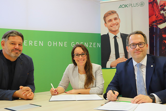 (v.l.n.r.) Mario Hanke und Linda Zölfel von der AOK Plus und Rektor Prof. Dr.-Ing. Alexander Kratzsch haben die Kooperationsvereinbarung vor sich auf dem Tisch liegen und lächeln in die Kamer.