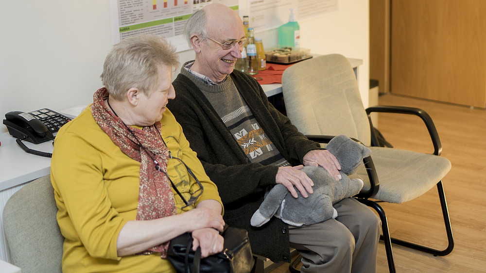Elderly couple has a stuffed cat on their lap and laughs.