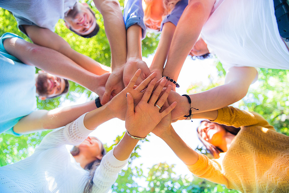 A group of people stand in a circle and place their outstretched hands on top of each other in the middle of the circle.