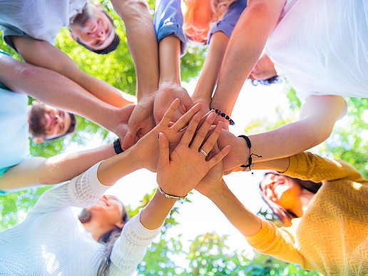 A group of people stand in a circle and place their outstretched hands on top of each other in the middle of the circle.