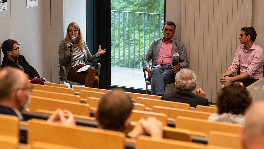 Four participants in a panel discussion are sitting in a semi-circle, discussing and listening to each other. Several participants are sitting in the auditorium.