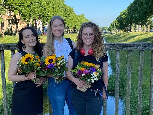 Three young women are standing on a bridge, smiling at the camera and holding bouquets of flowers that they have received for their German-Russian double degree...