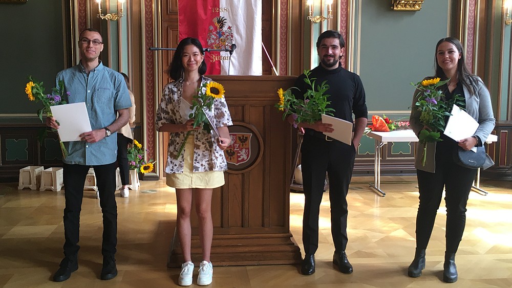 Vier Studienkolleg-Absolventinnen und Absolventen stehen im Bürgersaal des Zittauer Rathauses und halten ihre Abschlussurkunden in der Hand. Sie lächeln bei der Zeugnisübergabe in die Kamera.