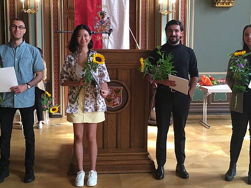 Four Studienkolleg graduates stand in the citizens' hall of Zittau town hall holding their graduation certificates. They smile into the camera as they receive their certificates.